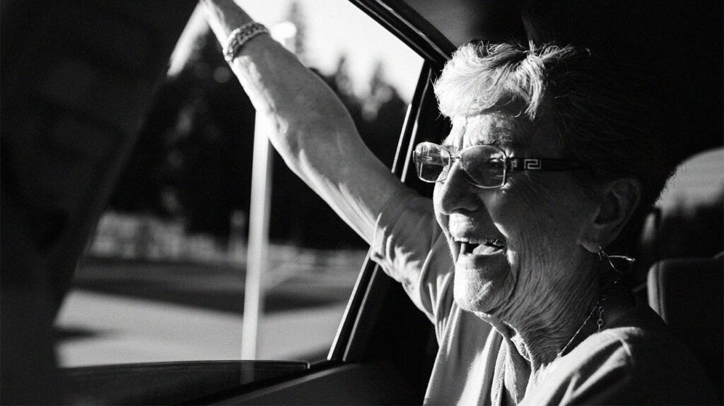 black and white photo of a smiling older woman with glasses waving through a car window