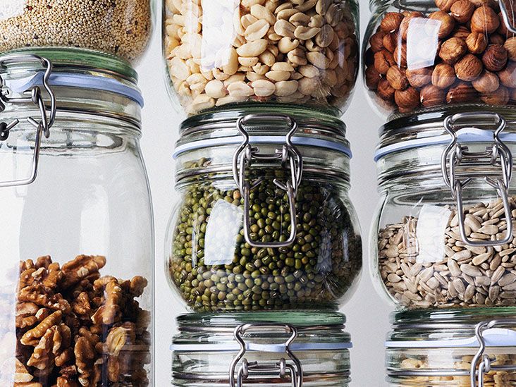 Several clear glass food storage containers are stacked one on top of the other and contain nuts, seeds, and legumes.