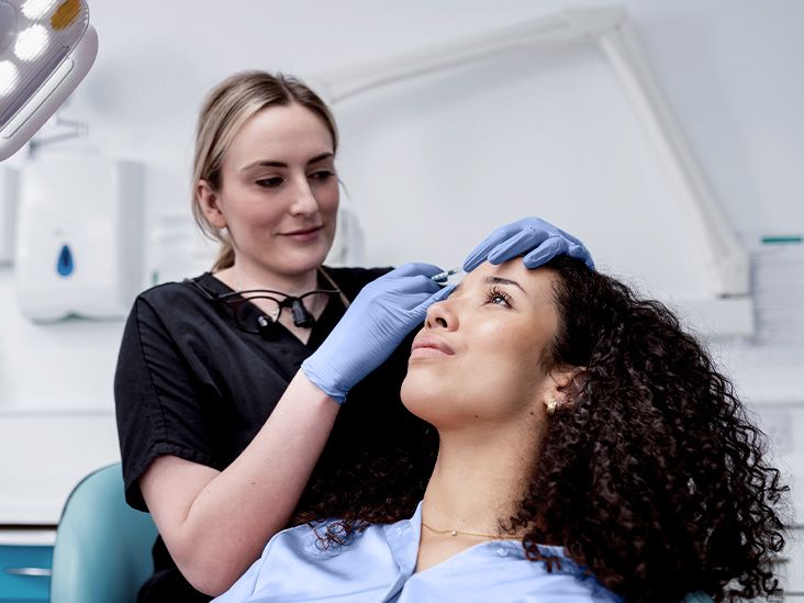 A women reclines in a doctor's office chair, waiting to receive botox treatments. A healthcare professional is next to her with a syringe of botox to administer into the women's forehead.