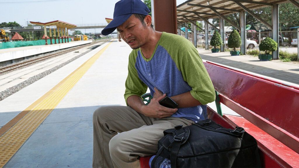 A person is sitting on a bench at a train station with their hands on their stomach.