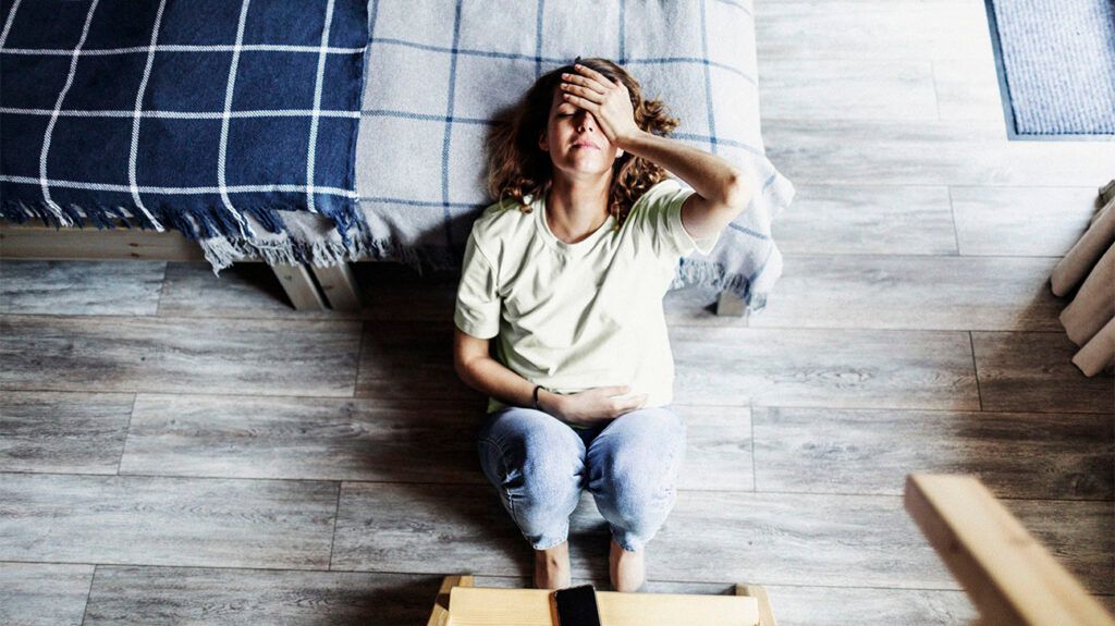 A woman is sitting on the floor, leaning against a bed, with her hand on her head.