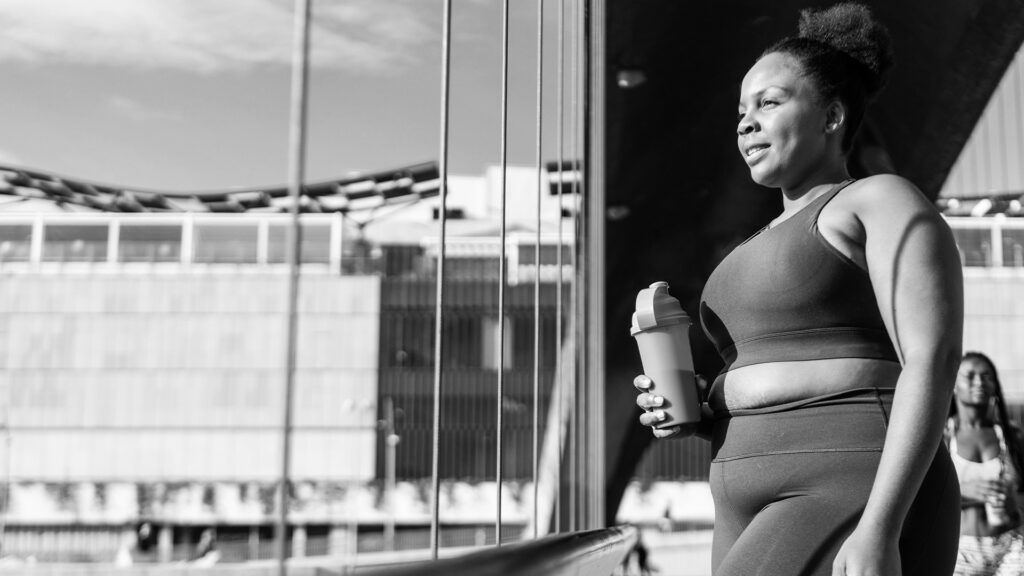a black woman in workout clothes looks out the window while holding a water bottle
