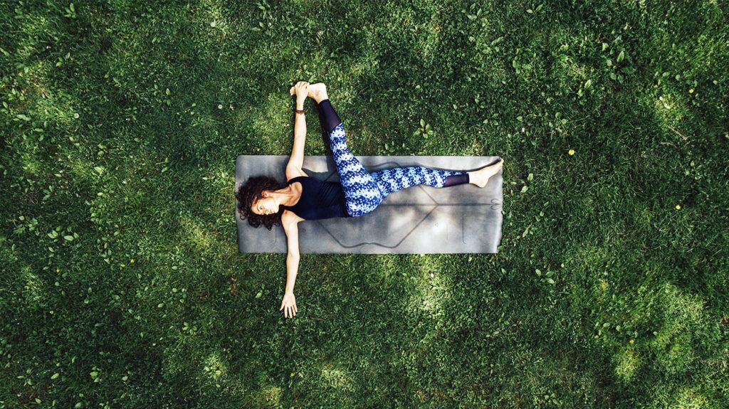 A woman doing a spinal twist yoga pose on a mat in a park, seen from above.