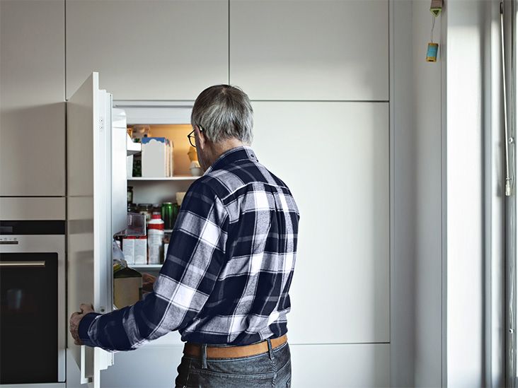 Man with gray hair and plaid shirt is opening a white refrigerator to examine the contents of food and medication in it.