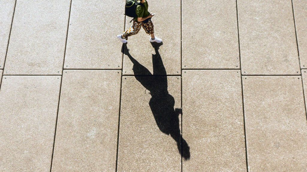 A person who may have oxidized cholesterol walking on a sunny pavement with a long shadow below them.-2