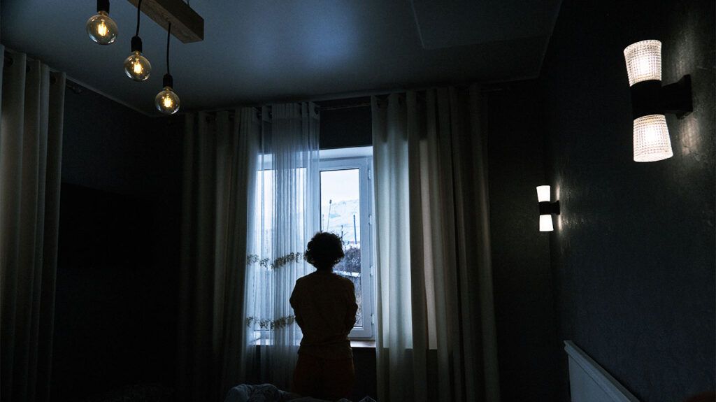 A person with migraine standing beside a window in a dark room to try and treat their symptoms.-2