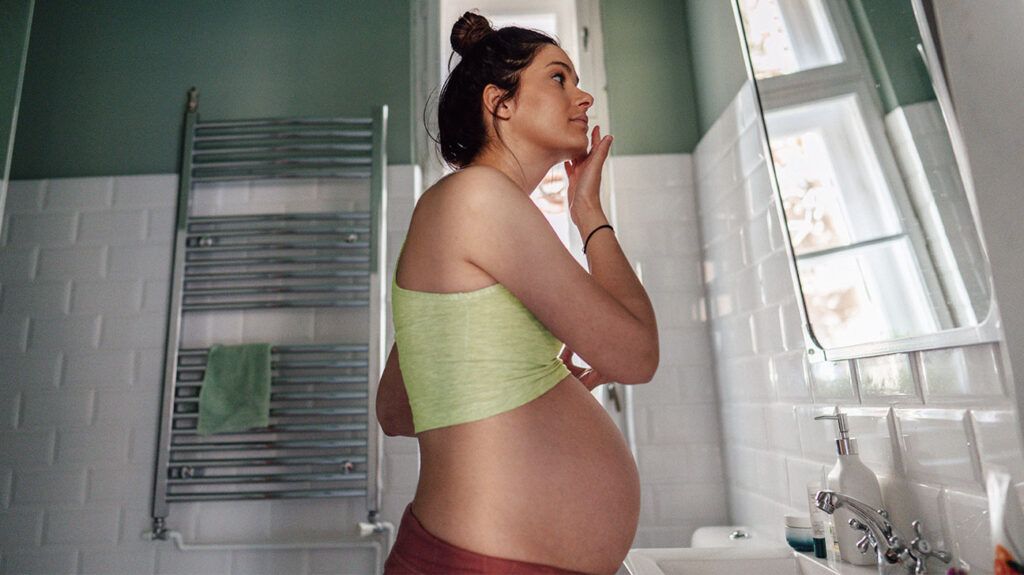 A pregnant woman applying face cream while looking in a bathroom mirror.