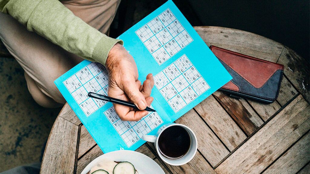 A hand holding a pen over sudoku puzzles on a wooden table.