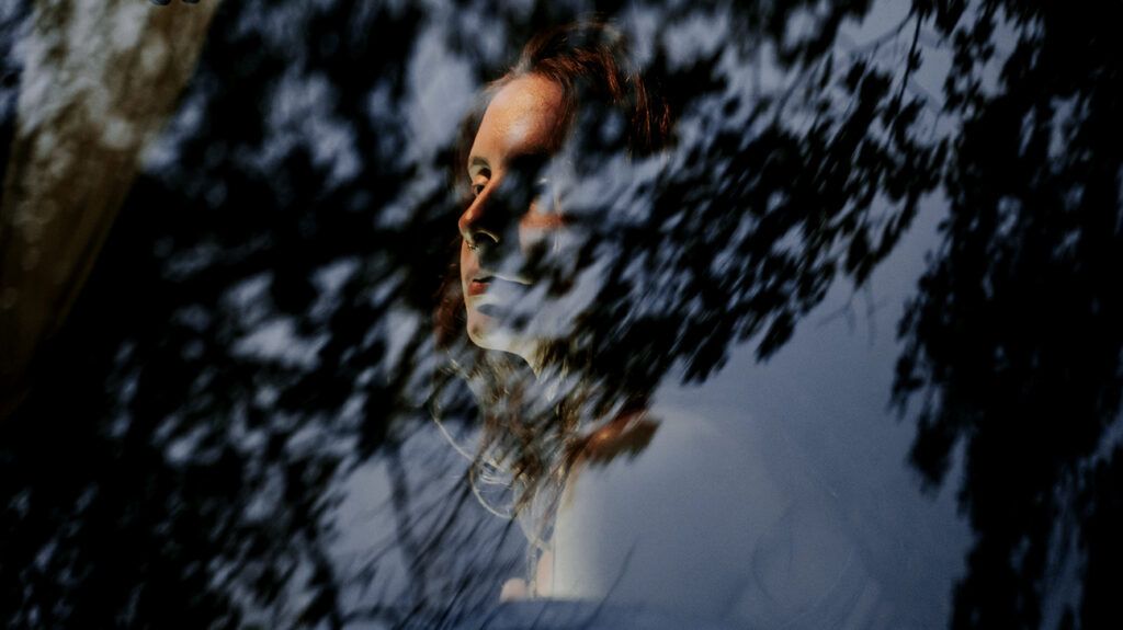 Photo of a woman with her face partially obscured by window reflections and shadows of leaves.