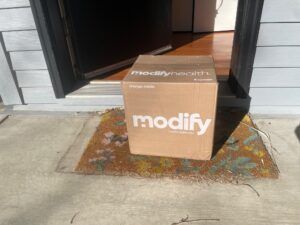 ModifyHealth meal kit delivery box sitting on welcome mat outside front door of a home