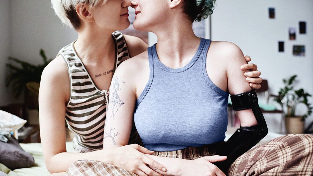 Two women embracing each other about to kiss. The woman on the right has a limb differences and wears a prosthetic device.