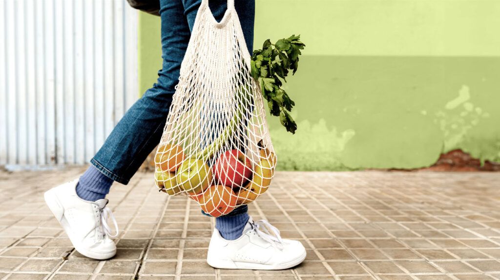 A person holding a bag of fresh fruits and vegetables walks on a sidewalk