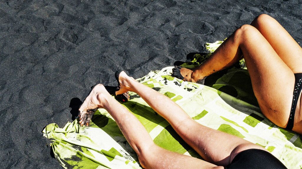 The legs of two people sunbathing on a beach with black sand.