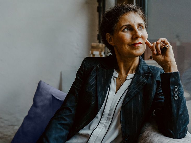 A women is dressed in a navy suit seated on a couch. She is looking out the with a slight smile on her face She looks happy..