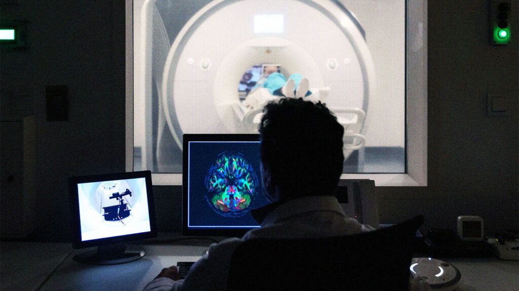 A person sitting in front of a computer to monitor a person undergoing an MRI scan. 