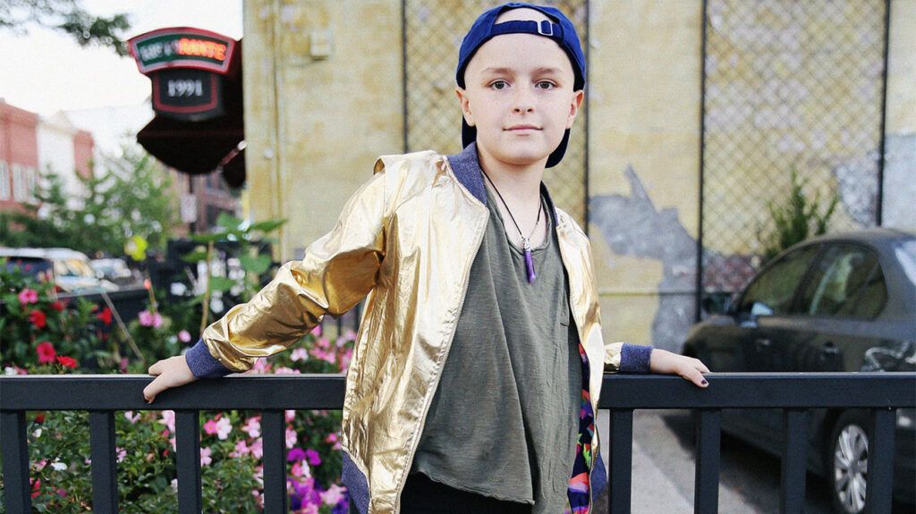 Younger child with Ewing Sarcoma wearing a gold jacket and standing in front of a metal fence