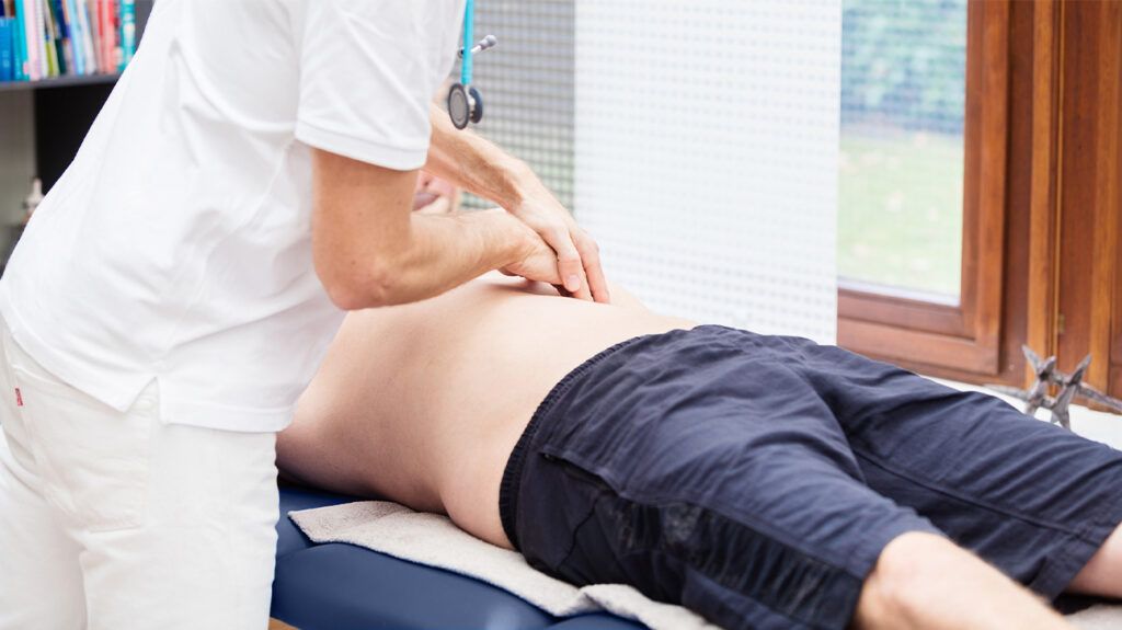 Doctor checking a person's abdominal mass. The patient is laying down in a medical setting, and the doctor is gently pressing on their abdomen