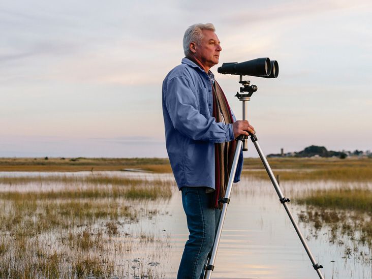 An adult male stands at the edge of a lake looking out into the landscape with a pair of binoculars.