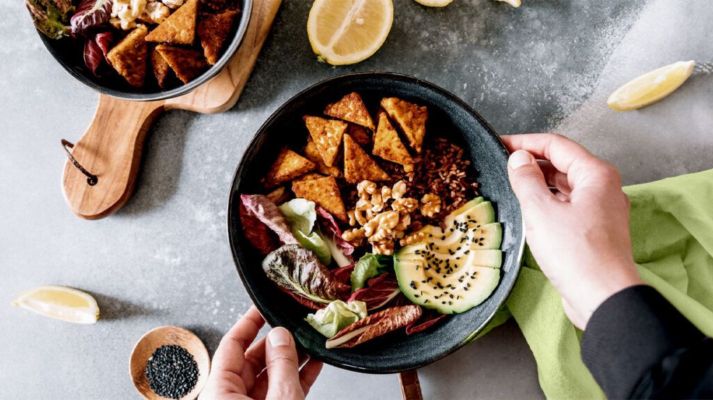 A vegan food bowl featuring greens, fried temphe, and avocado on a bed of grains on a table