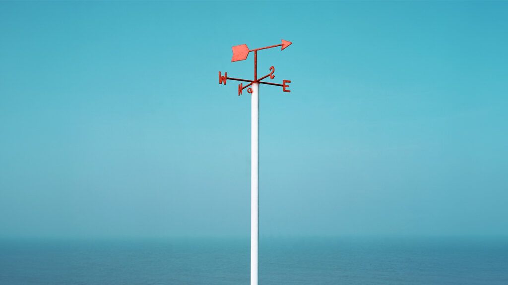 A weather vane pointing east with blue sky and sea in the background.