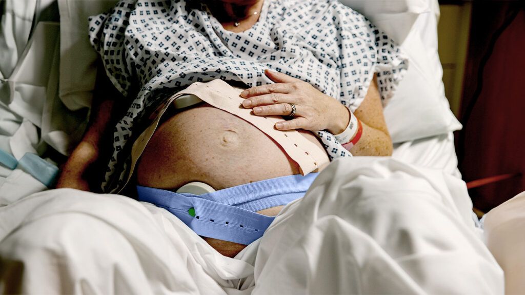 Torso of a pregnant person wearing a hospital gown and a heart monitor around the belly while sat in bed.