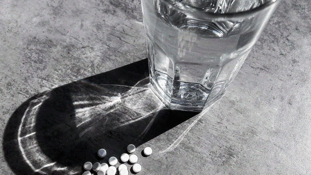 A handful of pills and a glass of water on a gray background