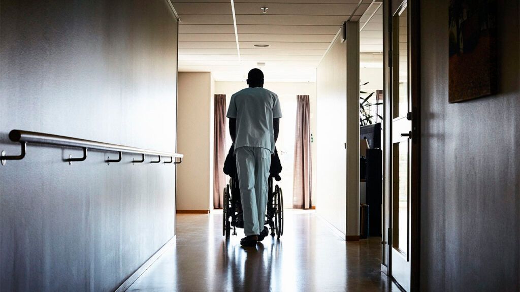 A medical professional is pushing a person in a wheelchair down a corridor.