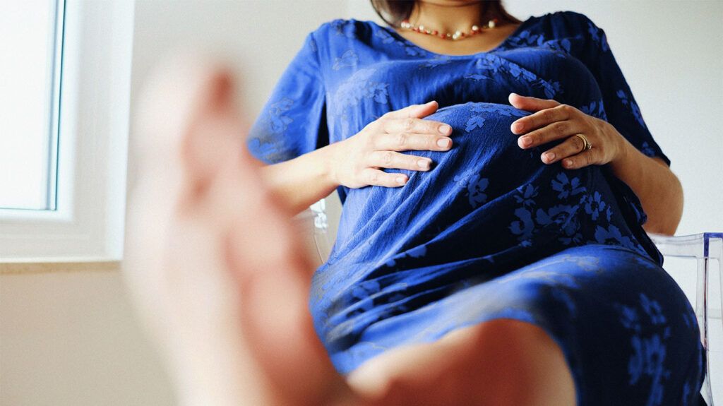 Torso of a pregnant person sitting in a hospital bed with hands on their bump.