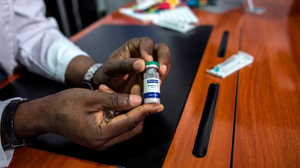 A person's hands holding a small bottle of glutathione. The person has a dark skin tone and is wearing a watch.