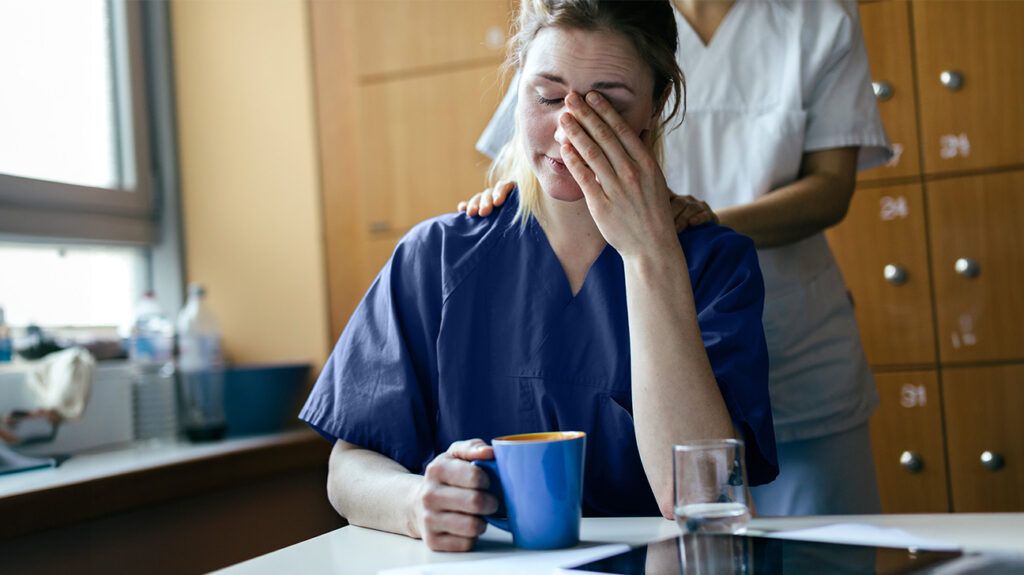 A tired healthcare worker with burnout drinking coffee while someone rubs her shoulders.