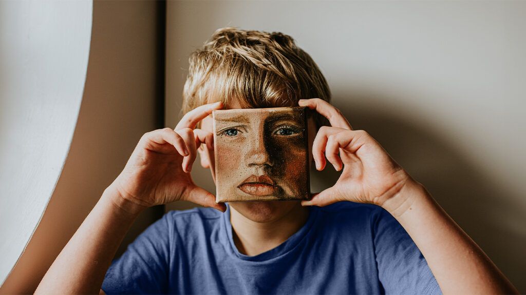 Young male child with a painting in front of his face of a sad face