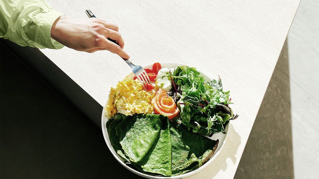 An overhead view of a person eating spinach pancakes, scrambled eggs, salmon, tomatoes and lettuce, ketogenic diet style