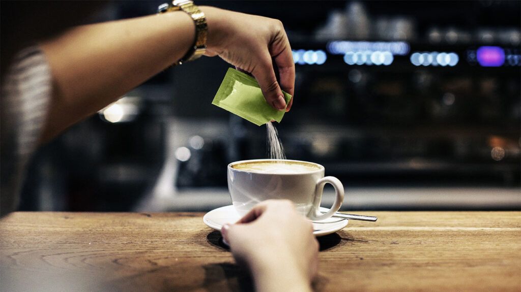 A person pouring a packet of sugar or sweetener into a cup of coffee