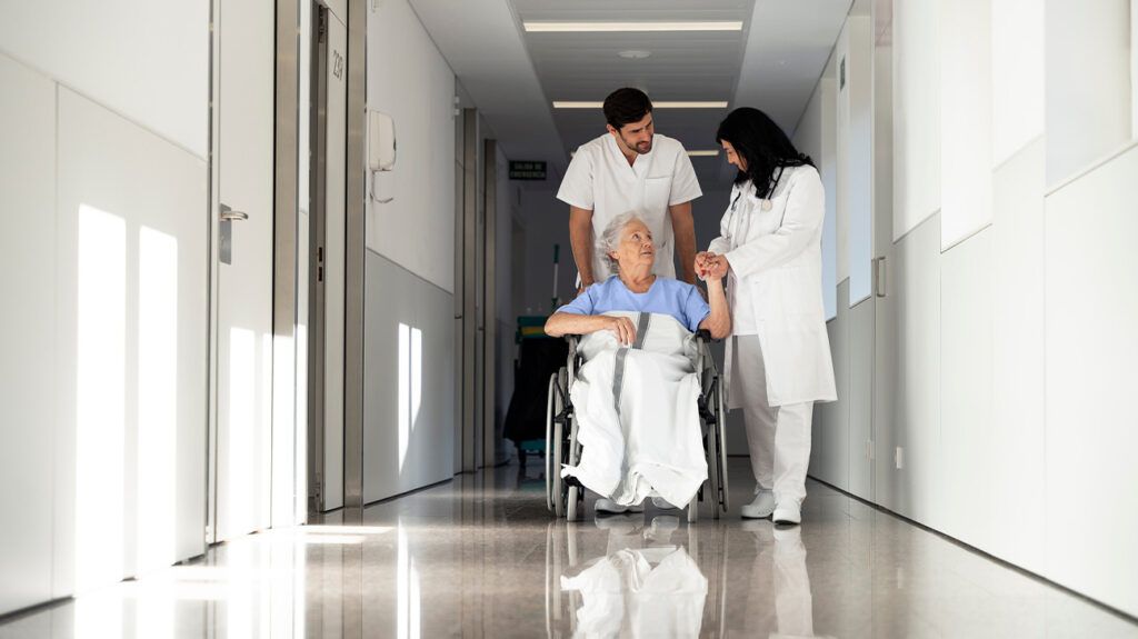 Older female sitting in a wheelchair in a hospital with two healthcare professionals