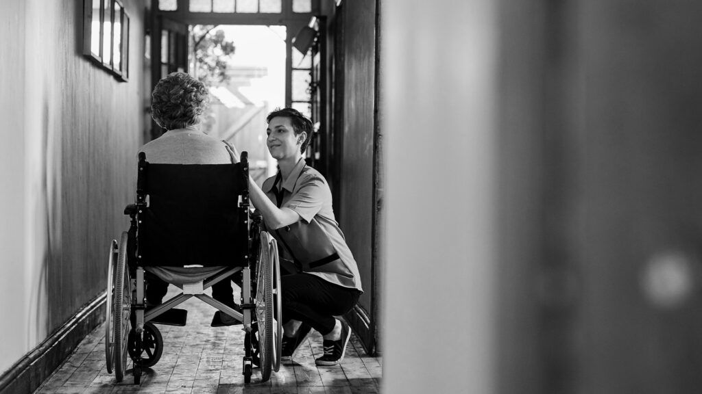 An older adult in a wheelchair, speaking with a nurse.