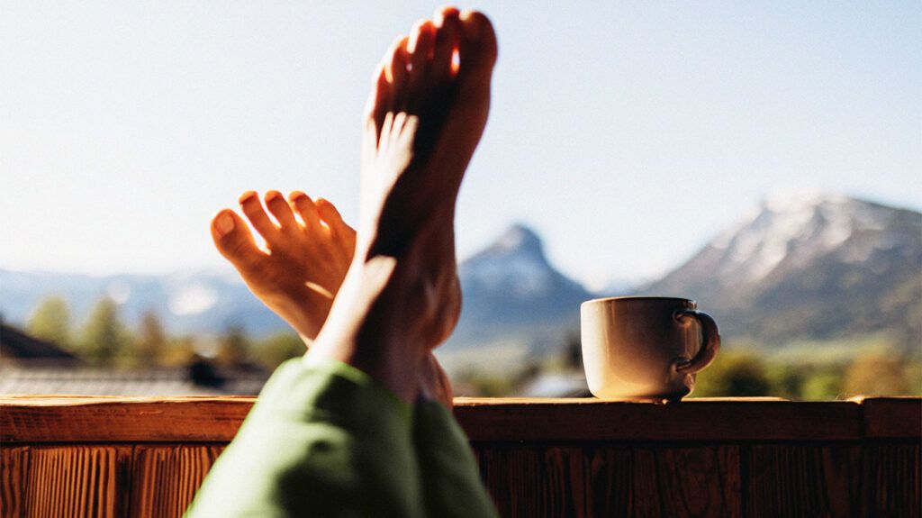 Person's legs and a morning cup of coffee against the backdrop of the Austrian mountains.