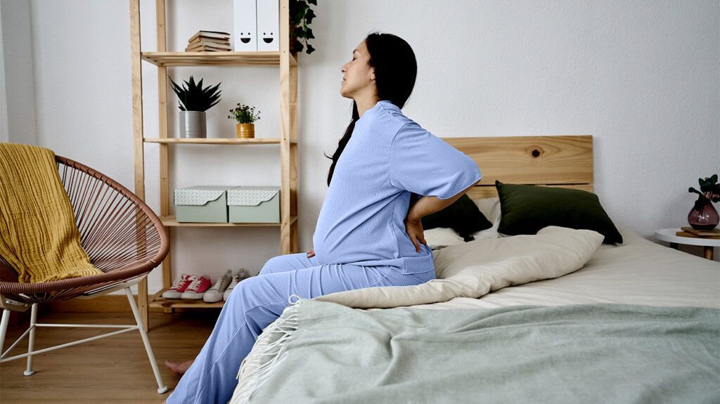 A pregnant person is sitting on a bed with their hands on their lower back.