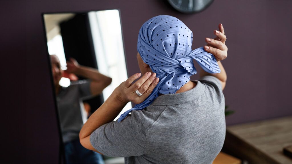 A woman undergoing chemotherapy for cervical cancer ties a headscarf around her head in front of a mirror