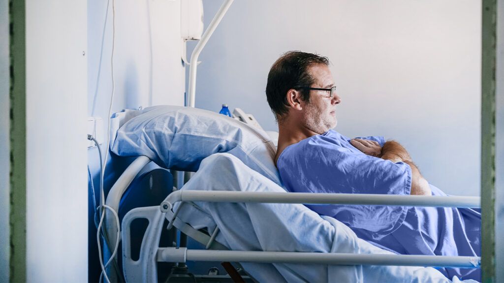 An older man sat in a hospital bed with crossed arms, seen in profile..