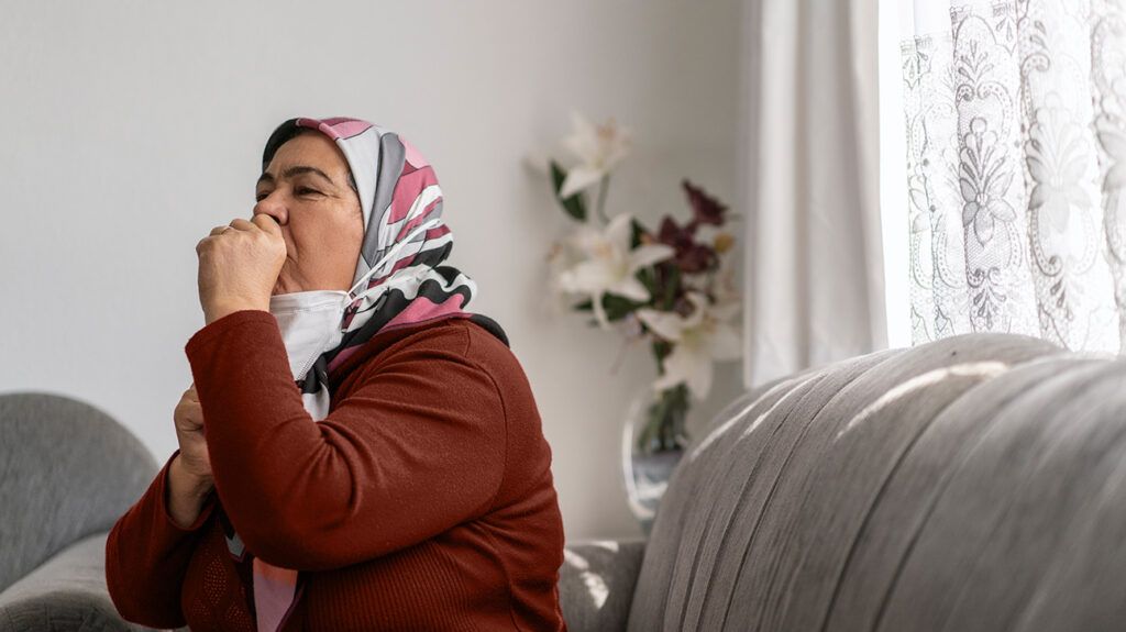 A woman coughing in a living room while sitting on a couch due to COVID rebound.