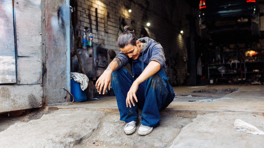 Une femme plus âgée est assise par terre devant un atelier automobile, fermant les yeux et posant sa tête sur un bras.