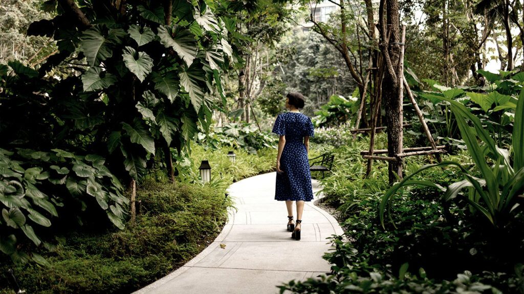 a man in a blue dress with white dots walks along the alley of a lush green park