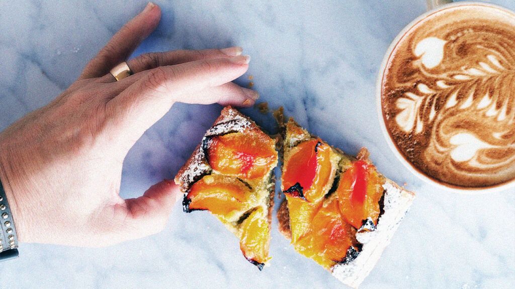 A person holding a piece of toast with fruit and a cup of coffee on a flat surface