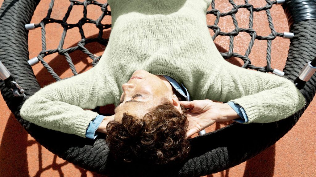 An older adult laying on a round rope swing at a playground, sleeping in the sun