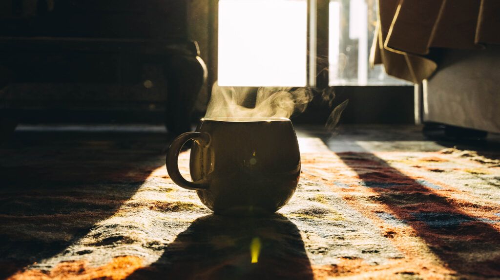 A mug of hot coffee on a carpet as light pours in from a window