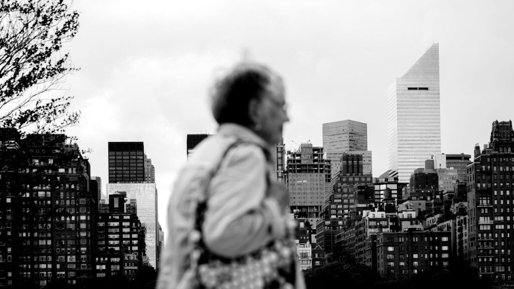 Black and white image of an older person walking in front of the New York City skyline