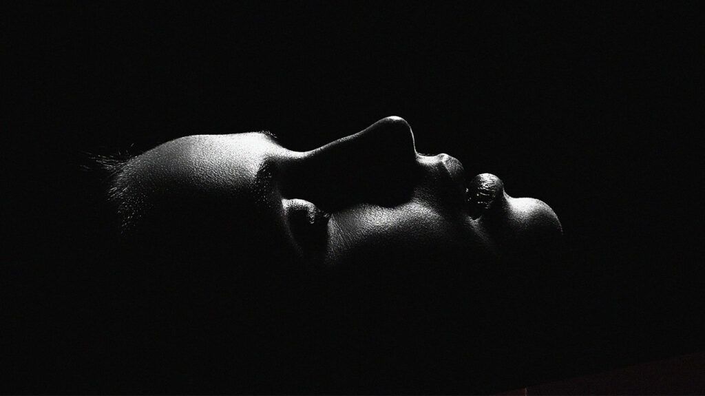 The profile of a woman's face in darkness with her eyes closed, facing up.