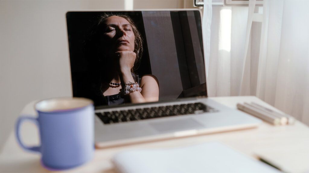 A reflection in a laptop screen of a woman with her eyes closed and chin resting on her hand.-1