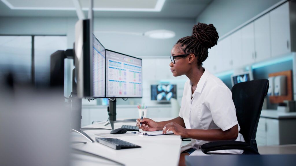 Adult female holding a pen and sitting at a double screen computer desk looking at billing and spreadsheets with x-rays and health information in the background
