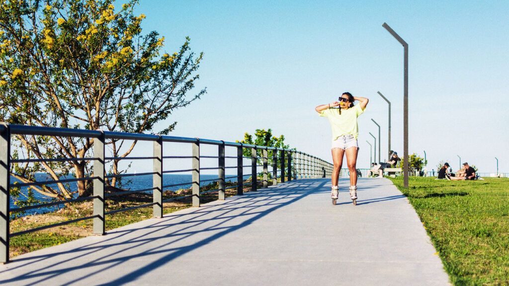 A person on rollerskates outside on a summer day's, while eating an ice cream and fixing their hair.